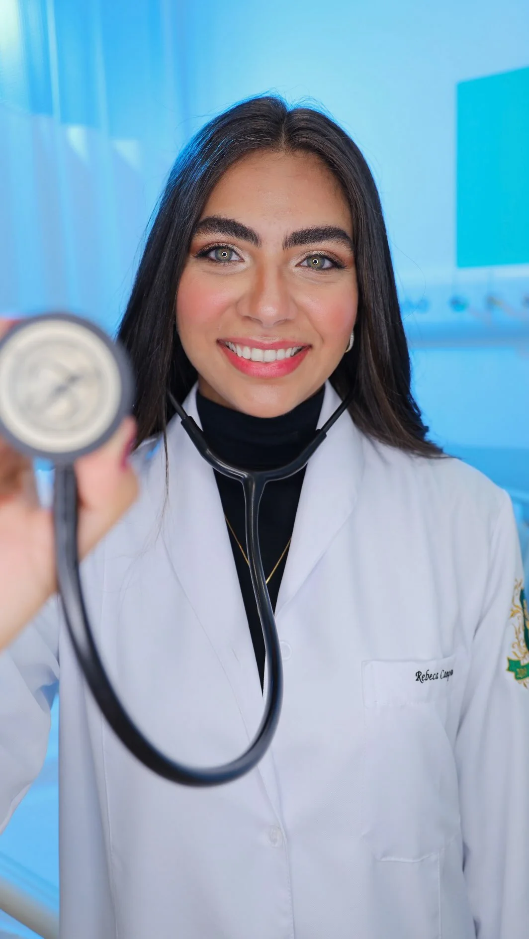 Rebeca Campano de jaleco branco, sorrindo e segurando um estetoscópio